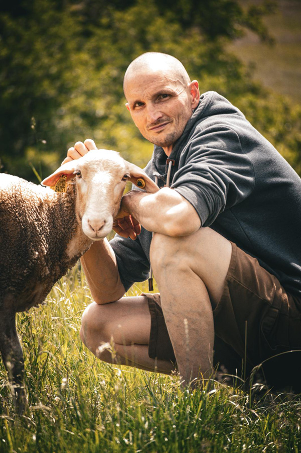 Olivier Monnet caresse un mouton tous deux le regarde tournés vers l'objectif au milieu des prés de Montpréval à Saint André sur Vieux Jonc en Terre de Rêves